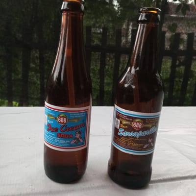 Two brown glass bottles, each containing a dark liquid and featuring a distinct blue-and-white label, are positioned upright on a white, textured surface. The scene is set outdoors in Teupinjok, Indonesia, with a soft-focus green background of foliage and indistinct dark structures.
The bottle on the left, identified by its label as "Rasa Ice Cream Soda," belongs to the "688" brand. Its label specifies a net content of 280 ml, a registration number (Reg. IDM 000109327), and indicates it was produced by "Perusahaan Lemon 'Asia Mon 688'" in Lhokseumawe, NAD, Indonesia. The label also displays a cartoon image of a girl drinking from a straw and an expiration date of "EXP. DECEMBER 2007."
The bottle on the right, also from the "688" brand, is labeled "Rasa Sarsaparilla." Its label includes a "HALAL" certification logo, a DIN. KES. P-IRT number (113117413 0022-25), and notes it as a "MINUMAN BERSODA BERKALORI RENDAH" (low-calorie carbonated drink). A cartoon image of a boy drinking from a glass is present, and like its counterpart, it states an expiration date of "EXP. DECEMBER 2007."
Both bottles exhibit consistent branding and production information. The white surface they rest upon shows slight creases. The background consists of blurry green vegetation, likely trees, providing depth to the image, with darker, out-of-focus vertical elements suggesting a distant fence or building structure. Natural light illuminates the bottles from the front-right.