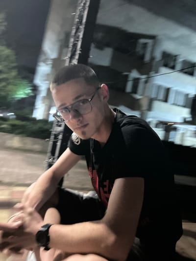 The image shows a young man wearing glasses and a black t-shirt. He is sitting outdoors at night, with buildings visible in the background. The man appears to be looking at the camera. The scene is dimly lit, suggesting it is nighttime. No specific landmarks identifying Dulovo, Bulgaria, are visible in the image.
