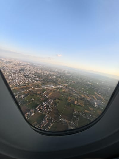 This is an aerial photograph taken from an airplane window, looking out over the city of Casalnuovo di Napoli, Italy. The perspective is from a high altitude, with the city sprawling across the landscape below. The image captures a mix of urban and rural areas, with residential buildings, agricultural fields, and infrastructure such as roads and what appears to be a train station. The sky is a clear blue with a few wispy clouds, and the sunlight suggests it is either early morning or late afternoon, casting long shadows across the terrain. The window frame of the airplane is visible along the bottom and left edges of the photograph, framing the view of the city and surrounding countryside.