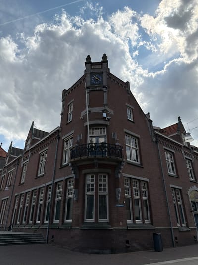 The image shows a multi-story brick building with white-framed windows and a distinctive clock tower. The building is located in Roermond, Netherlands, specifically at the intersection of Markt and Kloosterwandstraat. The architectural style suggests it is a historical building, possibly a public institution or a former guild hall. The sky is partly cloudy, with the sun visible through some breaks in the clouds. The street in front of the building is paved with cobblestones. There are no people visible in the image.