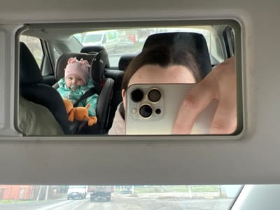 The image displays a reflection within an overhead rearview mirror positioned inside a vehicle, with the stated location being the city of Leninavan, Russia. The upper portion of a person's head, including the forehead and dark hair, is visible on the right side of the mirror's reflection. This individual is holding a light-colored smartphone horizontally, obscuring their lower face, with the device's three circular camera lenses clearly visible. To the left, a baby is seated in a dark-colored child safety seat, secured by straps. The baby wears a light pink hat with decorative ruffles and a blue-green patterned jacket, and has a pacifier in its mouth. An orange plush toy rests on the baby's lap. The external environment, reflected in the lower section of the car's windshield beneath the mirror, shows a paved road with vehicles, including a white van and a dark truck, and indistinct structures and vegetation in the background under an overcast sky.