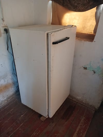 In a dimly lit room, a white refrigerator stands against a textured, off-white wall. The refrigerator, showing signs of age and wear, has a black handle on its door. To the left of the refrigerator, an electrical outlet with a black plug and cord is visible on the wall. The floor is made of dark wooden planks, some of which appear worn and stained. In the background, a window or opening emits a warm, yellow light, casting shadows on the wall. The overall impression is one of a rustic or possibly abandoned interior space. The scene is located in the City of Türkmenabat, Turkmenistan.