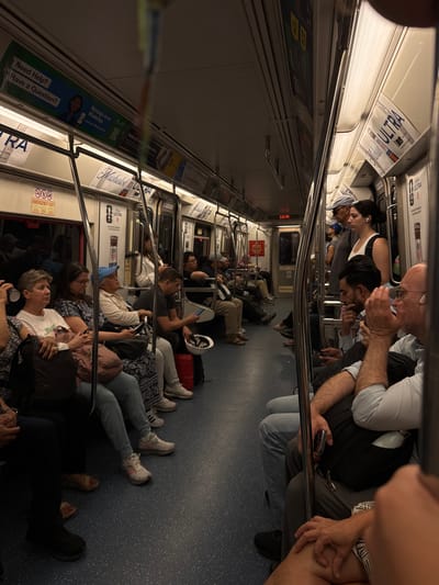 The media file captures the interior of a crowded train or subway car, likely in Jersey City, United States of America. The perspective is from a slightly elevated position, looking down the length of the car, with the image oriented to show people sitting and standing in a conventional train setting (the original file was rotated 90 degrees).
Numerous passengers of diverse ages are present. While some occupy the built-in seats, a significant number are seated or even lying down on the patterned blue floor in the aisle, suggesting crowded conditions, a prolonged journey, or a delay. Most individuals are seen looking at or interacting with their mobile phones, a common activity during travel. Some passengers are holding onto the overhead grab bars for support.
The setting is distinctly a train car interior, featuring seats, overhead luggage racks, and a series of advertisements along the upper walls. Other visible objects include personal items like backpacks, a red suitcase, a white hard hat, and various phones.
A digital clock mounted on the wall displays "3:33 PM." Advertisements for "ULTRA" and "Michelob" are prominent. Other visible text includes "Need Help? Have a Question? Message us on WhatsApp" with the associated URL "parynj.gov/WhatsApp," indicating a connection to the Port Authority of New York and New Jersey. Signs for "Reduced fares for eligible riders" and "EMERGENCY EXIT" are also present. The mood appears generally relaxed, with people engaged in their devices, comfortable in the crowded environment.