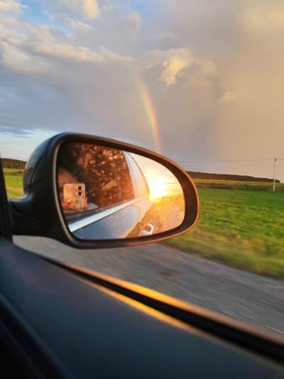 A car's side mirror reflects a sunset with a faint rainbow. The mirror also shows the face of a person holding a smartphone, taking a selfie. Outside the car, a green field stretches to a treeline under a cloudy sky tinged with the colors of sunset. The scene appears to be on a rural road, possibly near Bob'ya-Ucha, Russia, given the landscape and the rural feel. The image captures a moment of travel during sunset, with the natural beauty of the sky reflected in the car's mirror and the technology of a smartphone.