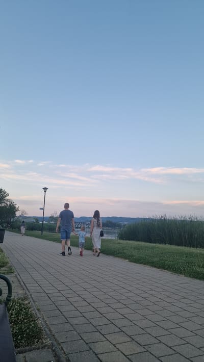 A family of three, a man, a woman, and a young child, are walking along a paved walkway next to a body of water. The man is wearing a blue t-shirt and denim shorts, and the woman is wearing a long, light-colored dress. The child is wearing a blue shirt and gray shorts. They are walking away from the camera, with the man holding the child's hand. In the background, there is a line of tall green reeds along the water's edge, and beyond that, rolling hills. The sky is a clear blue with wispy pink clouds. The scene appears to be set in a park or promenade, likely near the shore of Lake Balaton in Keszthely, Hungary, given the vegetation and body of water. A street lamp and a sign indicating "WC" are visible to the left of the walkway. Another person is walking further down the path on the left.