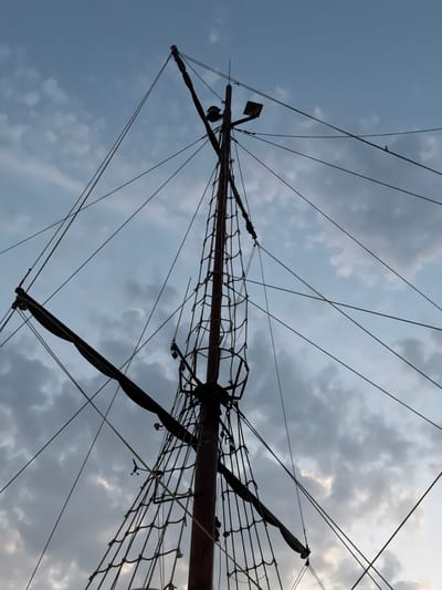 This is a low-angle shot of a ship's mast and rigging against a cloudy sky, likely taken in Sopot, Poland. The mast is dark and appears to be made of wood. Numerous ropes and lines crisscross the frame, connecting to various points on the mast and rigging. A furled sail is visible on the left side of the image. The sky is a blend of blue and gray clouds. No people are visible in the image.