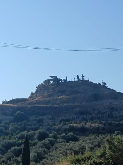 The image shows a landscape in the Municipal Unit of Evrostina, Greece. The foreground is filled with dense, dark green foliage, with a tall, slender cypress tree on the left side. Behind the foliage, a hillside rises, covered in dry, sparse vegetation and a few scattered trees. At the crest of the hill, a large, dark cross is silhouetted against a clear blue sky. Several smaller trees and what appear to be thin poles are also visible on the hilltop, clustered around the cross. Three thin, dark utility lines stretch horizontally across the upper portion of the sky.