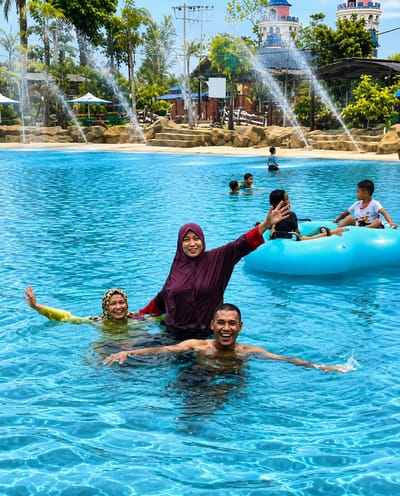 Four people are in a swimming pool at a water park, with water features and buildings in the background. In the foreground, a man and two women are swimming. The man is in the center, shirtless and smiling, with his arms outstretched. To his left, a woman in a yellow swimsuit and headscarf is also smiling with her arms out. Behind them, a woman in a dark purple burkini is waving. To the right of the frame, a blue inflatable raft is in the water with two children on it, one in a white shirt and the other in a dark shirt. The water is clear blue, and the background features lush green trees, fountains, and the colorful turrets of a water park attraction. The overall scene depicts a lively and enjoyable day at the water park.