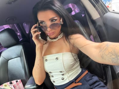 A woman with long dark hair is sitting in the driver's seat of a car, holding a smartphone to her ear and looking over her shoulder. She is wearing oversized sunglasses, a white strapless top with silver buttons, a pearl necklace, and dark pants with a brown belt. Her left arm, visible on her shoulder, has a pink constellation tattoo. The interior of the car is dark, with black leather seats. The exterior shows a hint of a city environment with buildings and graffiti visible through the car windows, suggesting a location within Alexandrovka, Russia. The overall scene captures a moment of a woman engaged in a phone call while seated in her vehicle.