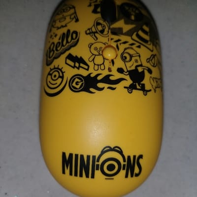 A yellow, elongated, capsule-shaped object, possibly a toy or gadget, is centered and depicted from an overhead perspective, resting on a light gray, faintly speckled surface. The object's upper surface is extensively covered with various black cartoon-style graphics from the "Minions" franchise. These include the stylized word "Bello," a Minion character skateboarding, a megaphone, a teddy bear, a traffic cone, lightning bolts, flame designs, the "Gru" (G) logo, and multiple Minion eye/goggle symbols. Near the top center, a small, spherical, light orange bead is suspended by a thin, dark rod or string. At the bottom of the object, the word "MINIONS" is printed in black capital letters, with a Minion eye graphic replacing the letter "O." The background surface is uniform in color and texture. There are no identifiable landmarks, signage, or environmental cues within the image to determine a specific location in 김포시 (Gimpo-si), South Korea.