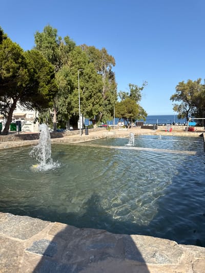 The image depicts a close-up view of a fountain with clear water cascading in several jets. The fountain is bordered by a stone wall on the right side of the frame, casting a shadow. Beyond the fountain, a body of water, likely the sea, is visible under a clear blue sky. The scene suggests a sunny day, possibly midday given the strong shadows.
In the background, lush green trees line a walkway or promenade. A few scattered objects, possibly benches or decorative elements like red flamingos, are visible along the waterfront. Small figures of people can be discerned in the distance, suggesting a public outdoor space. The architecture of some buildings is also visible behind the trees, hinting at a coastal town or resort.
The overall impression is one of a tranquil, sunny day in a pleasant, possibly Mediterranean, outdoor setting. The focus on the water and its movement creates a sense of refreshment and serenity. No discernible text is present in the image.