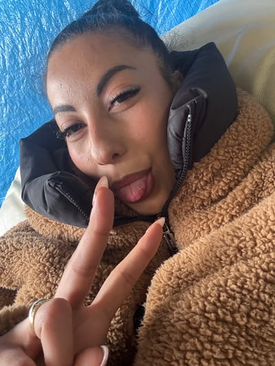 A woman is shown in a selfie, possibly indoors. She is lying on her back on a blue surface. The woman sticks out her tongue and gives a peace sign to the camera. She is wearing a brown fluffy coat with a black hood. The zipper is partly closed. The location of the photo is not available.