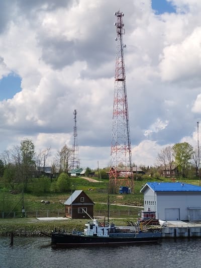 The image shows a waterfront scene in Ivanov Bor, Russia. A black and white tugboat is docked along a concrete pier. Behind the boat is a one-story, light gray building with a blue roof. To the left of the building is a small, dark brown wooden structure. Further back, the landscape rises gently with green grass, trees beginning to leaf, and several houses. Three radio towers dominate the background; one tall, red and white tower is prominent in the center, and two other towers are visible in the distance. The sky is partly cloudy with patches of blue visible.
