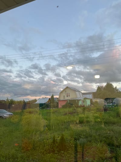 The image depicts a suburban scene in the village of Klyobushnevo, Russia, viewed through a reflective surface, likely a window. The foreground is dominated by tall, green grass and vegetation, with some orange debris visible in the lower left. A silver car is parked on the left side of the frame. In the mid-ground, several houses with pitched roofs are visible, along with fences and trees. One house is painted yellow with white trim, another is green with a silver roof, and a pink structure is partially visible on the far right. Several greenhouse structures are also present. The sky above is overcast with a mix of grey and white clouds, with hints of pink and pale blue suggesting either dawn or dusk. Three circular, illuminated fixtures are reflected in the viewing surface, appearing to be ceiling lights. Power lines are visible stretching across the upper portion of the image. The overall impression is one of a rural or semi-rural residential area during a transitional time of day.