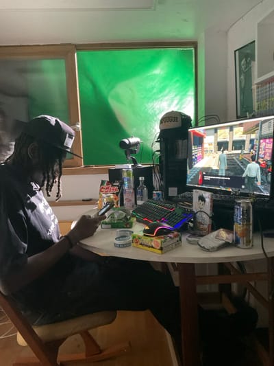 A person wearing a baseball cap and dark clothes sits at a cluttered desk. The desk holds a monitor displaying a video game, a keyboard, snacks, drinks, and a microphone. Behind the desk is a green screen. The person is holding a cell phone. The room appears to be dimly lit.