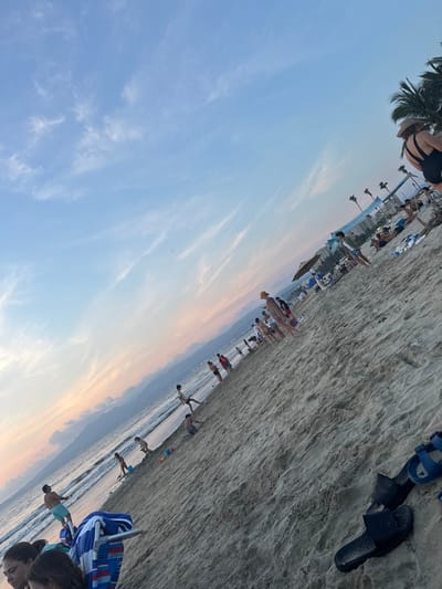 A beach scene is captured at an angle, revealing a sandy shore dotted with people and personal belongings like shoes. Palm trees sway in the background. The sky transitions from blue to a soft orange hue near the horizon, suggesting either sunrise or sunset. The ocean's edge is visible, with figures near the water, some appearing to be swimming. The image has a tilted perspective, with the sky dominating a large portion of the frame.