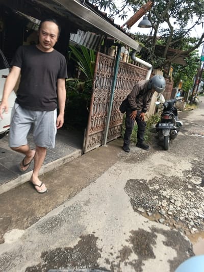 This image, captured in Cangkuang Kulon, Indonesia, depicts an outdoor scene featuring two adult males and a parked scooter.

On the left, an East Asian male with short dark hair, wearing a black short-sleeved t-shirt, grey athletic shorts, and dark flip-flops, stands partially emerged from a doorway or entrance. He is positioned with his left foot flat on the ground and his right heel raised, suggesting motion. He looks directly towards the camera. A white vehicle is partially visible inside the doorway behind him. Above the doorway, there is a corrugated metal awning with a white plastic rain gutter.

To the right, a second male, wearing a grey full-face helmet, a brown long-sleeved jacket, dark cargo pants, and black shoes, is stooped over. His back is to the camera, and his hands are placed on his knees as he bends forward at the waist. He carries a black shoulder bag slung across his body. Adjacent to him on the right is a black Honda Vario scooter, parked facing away from the camera, displaying license plate "D 6813 ON."

Between the two individuals is a rusty brown metal gate with an intricate, open scrollwork design, appearing partially open. Green foliage is visible behind the gate. Above the gate and the second male, a broken or detached section of a corrugated metal roof is visible, along with a dark-colored, bell-shaped hanging lamp attached to a white PVC pipe. The area behind the scooter shows a street or pathway with trees and utility infrastructure in the background.

The ground in the foreground is uneven concrete, marked by patches of moisture and scattered small stones or gravel. The overall lighting suggests an overcast day.