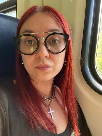 The image presents a headshot of a female individual with long, straight red hair, seated inside what appears to be a train or bus car. She has a medium skin tone and brown eyes, looking directly at the viewer with a neutral expression. The individual wears eyeglasses with prominent roundish black frames and a gold-colored metal bar extending across the top of the bridge. Green reflections are visible on the lenses, consistent with outdoor foliage seen through a window. She is adorned with silver-colored hoop earrings and a silver-colored chunky chain necklace featuring a cross pendant. Her attire consists of a dark-colored top, possibly black or dark gray, with a sheer, textured overlay. To the left, a portion of a blue upholstered seat is visible. To the right, a window with a black frame reveals a blurred outdoor scene primarily showing green foliage, indicating motion and daylight. The interior wall or ceiling paneling of the vehicle is a light, neutral color. The scene is located in the city of Vyatskiye Polyany, Russia.