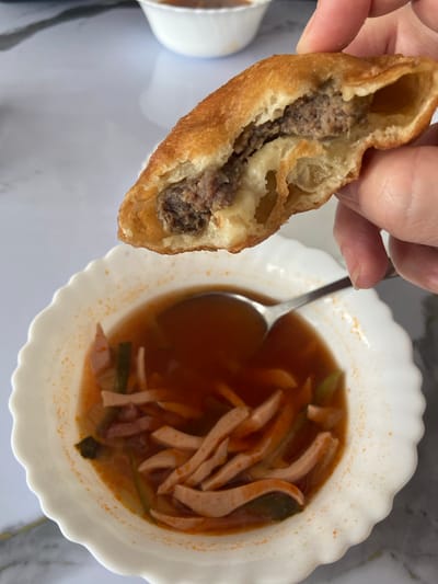 A hand is holding a half-eaten, fried pastry filled with ground meat and cheese over a white bowl of soup. The soup appears to be a broth with sliced sausage and some green vegetables. The background is a white, textured surface. There is no indication of the location being in Усмангали, Russia.