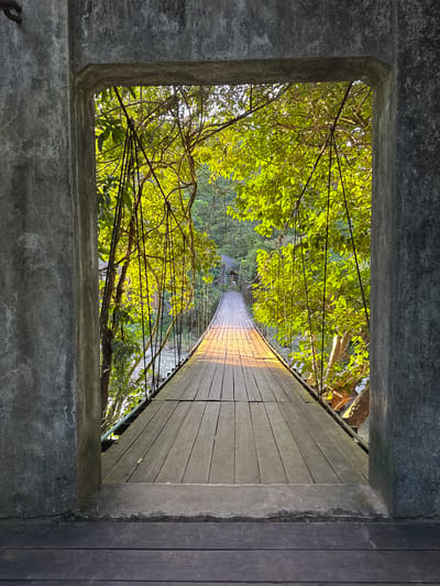 A wooden suspension bridge spans a river, seen through a concrete archway. The bridge is made of weathered wooden planks and is supported by cables. Lush green trees with bright sunlight filtering through their leaves line the sides of the bridge, creating a dappled effect on the wooden walkway. The river flows beneath the bridge, with hints of vegetation on the opposite bank. The scene is set in Ranong, Thailand, likely a natural attraction or park. There are no people visible in the image.