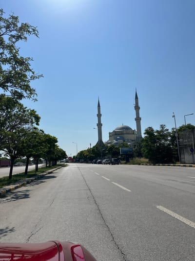 This image captures a wide-angle view of a multi-lane asphalt road on a clear, sunny day, with a prominent mosque in the mid-ground. The location is Edirne Merkez, Turkey, specifically facing the Darülhadis Mosque (Edirne Darülhadis Camii), identifiable by its distinct architecture featuring a large central dome flanked by smaller domes, and two tall, slender minarets with multiple balconies, constructed from light-colored stone or concrete. The sky is a cloudless, vibrant blue, indicating bright daylight.

In the foreground, the hood of a red vehicle, likely the one from which the photograph was taken, is visible at the bottom. The road surface exhibits numerous cracks and wear, with clear dashed white lines marking lanes and a solid white line on the right shoulder. A yellow and black striped curb runs along the right side of the road, bordering a narrow grassy verge and a sidewalk.

Trees with green foliage line both sides of the road, casting shadows on the left side, consistent with the sun being high in the sky. On the left side of the road, further down, several vehicles including a large red truck and a white caravan/trailer are visible in the distance. On the right side, closer to the mosque, a few parked passenger cars are visible behind the trees and a low concrete barrier.

Adjacent to the Darülhadis Mosque on the right, partially obscured by trees, are other structures including a tall pole with a security camera mounted at its top, a streetlight, a large billboard with an image that appears to be a landscape, and a concrete wall or fence. No people are visible or identifiable in the scene. The overall impression is one of a major urban thoroughfare with relatively light traffic, passing by a significant religious landmark.