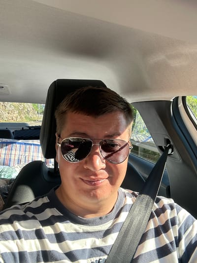 The image shows a close-up, sideways, and slightly angled selfie taken from inside a car. The primary subject is a person wearing sunglasses and a blue and white striped t-shirt. The person's face is partially visible, with their mouth and chin in focus, and their eye visible through the reflection on the sunglasses. The sunglasses have a metallic frame and dark lenses, reflecting the person's striped shirt and a pink object.

The setting is the interior of a car, evidenced by the car's interior paneling, a seatbelt, and a car seat. The seatbelt is prominently displayed across the person's chest and is clipped into its buckle. Sunlight is streaming into the car, creating bright highlights and shadows, suggesting it is daytime. Through the car window, a blurry view of greenery and what appears to be a sunny outdoor environment can be seen, indicating pleasant weather. Behind the person, a patterned blanket or fabric is visible on the car seat. The overall mood seems casual and relaxed, possibly suggesting the person is on a road trip or enjoying a sunny drive. The location context provided is Provodina, Montenegro. There is no visible text in the image.