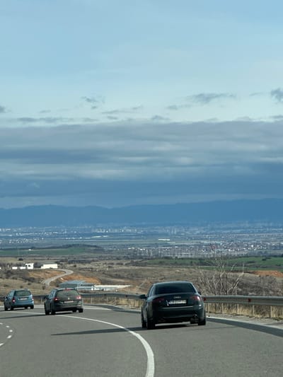 The image depicts a panoramic view from an elevated paved road in the city of Bogyovtsi, Bulgaria, looking northeastward towards a distant urban area, likely Sofia. The foreground features a two-lane asphalt road curving sharply to the right, marked by a solid white line on the inner edge and a dashed white line on the outer. Three vehicles are observed on this road, moving away from the viewer. The nearest vehicle is a black Audi sedan, bearing Bulgarian license plate CB 2099 PH. Ahead of it, a dark grey hatchback is visible, followed by a light blue station wagon further in the distance. A metal guardrail parallels the right side of the road.

The midground consists of a sprawling valley characterized by undulating terrain with patches of green and brown vegetation, consistent with agricultural fields and scrubland. Industrial structures and scattered white-roofed buildings are discernible across the landscape. The background features a large city with numerous buildings and structures, appearing somewhat hazy due to distance, situated at the base of a distinct, dark blue mountain range that spans the horizon. The sky overhead transitions from a clear, pale blue at the zenith to a band of layered, grey-blue stratus clouds occupying the middle and lower sky. No individuals are visible in the scene. The environment appears to be an overcast day with cool weather.