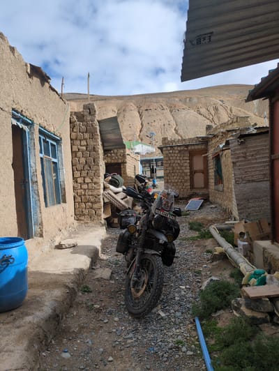 The image depicts a view of a village in Pang, India, with a motorcycle parked in the foreground. The village consists of structures made of mud and stone, with corrugated metal roofs. A blue plastic barrel is visible to the left of the frame, and a motorcycle, laden with luggage, is positioned centrally. The ground is covered in gravel. In the background, a barren, mountainous landscape is visible under a cloudy sky.