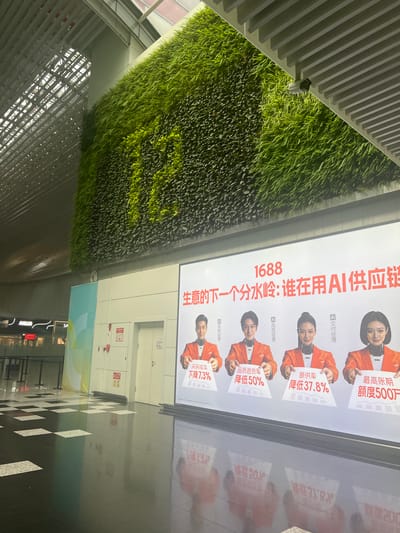 The image shows a large, modern interior space with a prominent vertical green wall on the left and a large digital advertisement billboard on the right. The space appears to be a public area, possibly an airport, train station, or shopping mall, with a high ceiling and a sleek, metallic ceiling structure. The floor is a checkered pattern of black and white tiles, with a highly reflective surface that mirrors the surroundings.
The green wall is lush and vibrant, covering a significant portion of the background on the left side. The billboard on the right displays a vibrant advertisement with the number "1688" in large red font at the top. Below this, there is Chinese text, which translates to "The next watershed moment for business: Who uses AI in the supply chain?". The advertisement features four individuals, two men and two women, all wearing orange jackets with AI-themed details around their necks. Each person is holding a sign with Chinese text indicating a business metric and a percentage reduction. For example, one sign reads "Purchase conversion rate reduced by 7.3%". The people appear to be models or actors, and their faces are digitally enhanced to have a somewhat artificial or AI-like appearance.
In the background, to the left of the green wall and behind a railing, there are signs for shops, including "AIGLE", indicating a retail environment. There are also white doors and panels that look like lockers or service access points on the wall behind the billboard. The overall impression is one of a clean, contemporary, and technologically advanced public space. The lighting is bright and diffused, suggesting an indoor environment with artificial illumination. The image has a slight tilt and is taken from a medium distance. There are no people visible in the immediate foreground or interacting with the advertisement. The reflection on the floor indicates the presence of the advertisement and the ceiling structure.