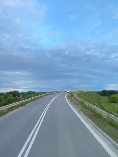 The image shows a two-lane asphalt road with white markings, curving to the right. The road is bordered by a metal guardrail on the left and a grassy embankment with a guardrail on the right. Green trees and vegetation are visible on both sides of the road, with some distant buildings and structures. The sky is cloudy and appears to be late afternoon or early evening. A white car is visible in the distance on the road. Based on the provided information, this scene is located in Podłęże, Poland.