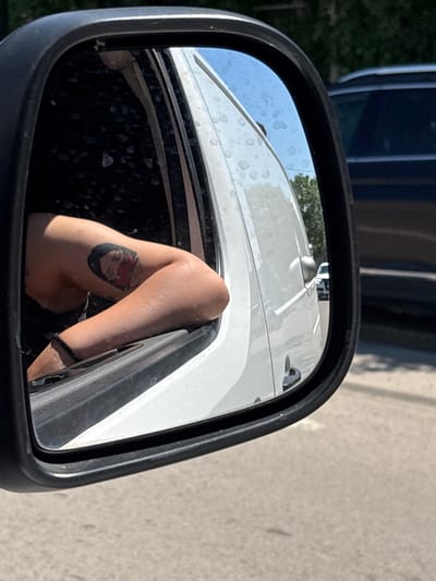 The image is a close-up, distorted reflection in a side-view mirror of a vehicle. The primary subject of the reflection is a person's legs, with one leg bent at the knee and the other extended. A tattoo is visible on the calf of the bent leg, depicting a circular design with a red element and a black outline. The person appears to be wearing a dark band around their ankle.
The reflection also shows parts of a white vehicle, likely a boat or RV, with some water spots visible on its surface. The background of the reflection includes a blue sky and some greenery, suggesting an outdoor setting. The mirror itself is framed by a black casing, and a portion of the vehicle's interior, which appears dark and possibly dirty, is visible in the lower part of the mirror.
The scene is brightly lit, indicating it is daytime, likely sunny given the strong reflections and shadows. The overall impression is casual and personal, possibly taken by the person themselves. The location context of Constanța, Romania, does not offer specific visual cues within the image itself, but suggests a coastal or lakeside environment. No text is visible in the image.