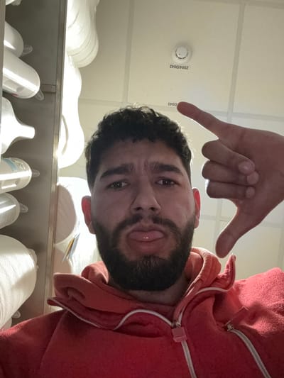 A male individual is positioned centrally, looking directly at the camera. He has dark, curly hair and a full dark beard. He is wearing a red hooded sweatshirt with a white zipper. His tongue is slightly extended. His right hand is raised on the upper right side of the frame, with the index finger and thumb extended upwards, and the other fingers curled, forming a downward 'L' shape. To the left of the individual, several large, white, cylindrical spools or rolls, possibly industrial thread or fabric, are stacked vertically on shelving. A bright light source illuminates these objects from the left. The ceiling is white with a grid pattern, featuring a circular smoke detector labeled "Z004/D042" and an adjacent label "D043". The setting is an interior industrial facility, warehouse, or storage area, characterized by its functional infrastructure and stored materials, located within the city of Radovljica, Slovenia.
