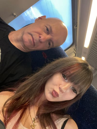 The image is a selfie taken inside a train. The foreground shows two individuals. On the right, a young woman with long, dark brown hair with red highlights, bangs, and dark eye makeup poses with her lips puckered. She is wearing a white top and a gold necklace with a heart and an angel pendant. On the left, an older bald man with fair skin is partially visible, his head tilted towards the woman. He is wearing a black t-shirt. The background shows the interior of a train carriage, including blue seats, a window with a view of a bright sky, and fluorescent lighting. The image was taken in the city of Azor, Israel.