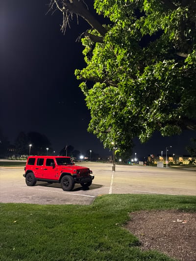 In an outdoor parking lot at night in Maryland Heights, Missouri, a red four-door Jeep Wrangler is parked. The vehicle is positioned in the foreground, angled slightly towards the viewer, with its front end visible. The Jeep has black tires and a black grille. To the right of the Jeep, a large tree with green leaves dominates the frame, its branches extending overhead and partially obscuring the dark night sky. The parking lot is paved and marked with white lines. In the background, streetlights illuminate the lot, and several buildings with lighted windows are visible. A few faint white specks, possibly birds or insects, are scattered in the sky. The ground in the lower portion of the image is covered with green grass.