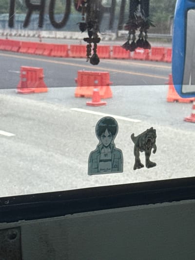 The image presents a view from inside a vehicle, looking outward through a window onto a road in Bang Sao Thong Subdistrict, Thailand. Two stickers are adhered to the lower section of the window: one is a light-toned depiction of an anime character, identified as Eren Yeager, facing forward with a neutral expression; the other, positioned to its right, is a three-dimensional figure resembling a roaring bipedal dinosaur or monster. Reflections are visible in the upper part of the window, including mirrored text partially legible as "IMPORT" and dark, ornate hanging items, possibly vehicle decorations. A portion of a blue vehicle component, likely a side mirror housing, is visible on the extreme right. The exterior environment consists of an asphalt road with visible white lane markings. Numerous orange plastic road barriers and red traffic cones are arranged along the road, indicating a controlled or work zone.