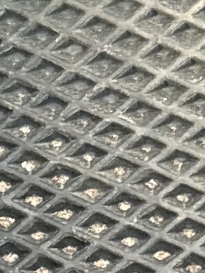 The image shows a close-up of a textured surface, likely a material with a repeating pattern of raised diamonds. The surface appears to be dark gray, with some lighter-colored specks scattered across it. The texture is rough and three-dimensional, with distinct shadows and highlights indicating depth. The pattern is uniform and covers the entire visible area of the image. There are no people, animals, or any discernible objects beyond the texture itself. The lighting appears to be neutral, suggesting an indoor or overcast outdoor setting. There is no visible text in the image. The context provided, "Пашкаўскі сельскі Савет, Belarus," suggests a rural location in Belarus, but the image itself does not offer any specific visual cues to confirm this. The image is abstract in nature, focusing solely on the visual properties of the textured surface.