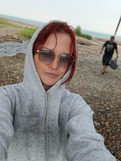 The image captures a selfie taken by a young woman with auburn hair and large sunglasses, wearing a grey hooded sweatshirt. She is positioned in the foreground, facing the camera with a slight smile. In the background, a man wearing a black t-shirt and shorts walks away from the camera, carrying a dark bag. The setting appears to be a beach with grey pebbles and a body of water in the distance, likely the Black Sea given the location of Priboi, Russia. The sky is overcast.