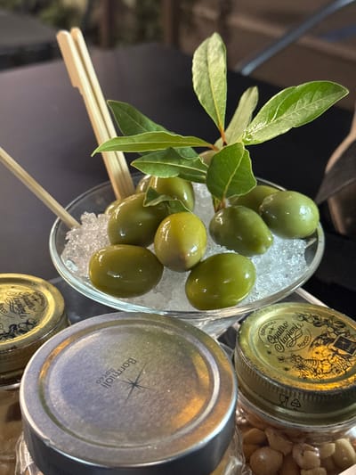 A close-up shot reveals green olives presented on a bed of crushed ice in a clear glass bowl. A sprig of bay leaves adorns the olives, adding a fresh touch. The bowl also has wooden skewers to pick the olives. Nearby are several jars with metallic lids, including one labelled "Bormioli Rocco" in black print. The jars appear to contain preserved food items. The composition is neatly arranged, offering a glimpse into an appetizer or snack preparation.