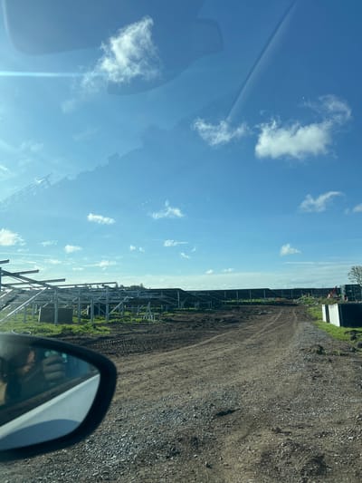 The image appears to be taken from inside a vehicle, possibly a car, with a view out towards what seems to be a construction site or a rural area. The sky is blue with some scattered clouds. A metal framework, likely for solar panels, is visible along the left side of the frame. A dirt or gravel road runs along the right side of the frame. In the distance, there are some structures that may be buildings or storage containers. The image includes part of the vehicle's side mirror in the lower right corner, with a blurred reflection of the driver or passenger visible. The overall impression is of a somewhat industrial or development area. The image does not definitively confirm the location is Grange Ville, Ireland.