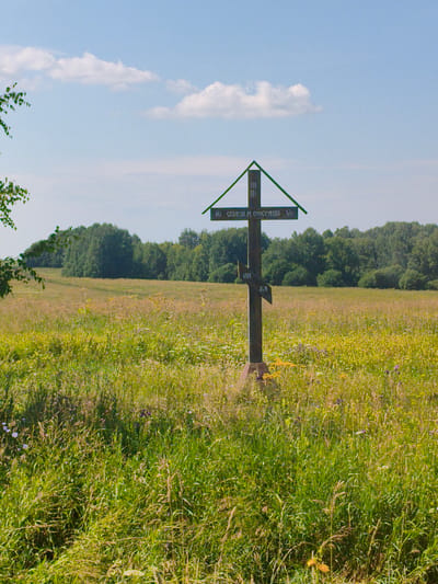 A wooden Orthodox cross stands in a grassy field under a blue sky with scattered white clouds. The cross is dark brown with a green slanted roof-like structure at the top. Cyrillic text is inscribed on the cross. The field is covered in tall green grass and wildflowers in various colors, including yellow and purple. In the background, a dense line of green trees borders the field. The lighting suggests it is daytime, likely midday or afternoon. The scene appears to be in a rural or natural setting, possibly the city of Bannovo, Russia, given the context. There are no people or animals visible in the image.