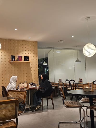 The image depicts two women wearing hijabs sitting at a table inside a cafe in Ngaglik, Indonesia. The woman on the left wears a white hijab and a plaid shirt, while the woman on the right wears a black hijab and a black jacket. They are seated opposite each other at a small, round table. The table has a pitcher of water, glasses, and some belongings on it, suggesting they are engaged in a conversation or having a meal.

The cafe's interior features a distinctive wall with a textured wooden paneling adorned with circular indentations. A shelf on this wall displays three dark-colored books. Other tables and chairs are visible in the background, some with woven seats and chrome frames. A large, round, white paper lantern hangs from the ceiling, providing ambient light. A glass partition separates this area from another section of the cafe, where more tables and chairs are set up. The flooring is tiled in a light gray. The overall atmosphere appears to be relaxed and casual, typical of a cafe environment.