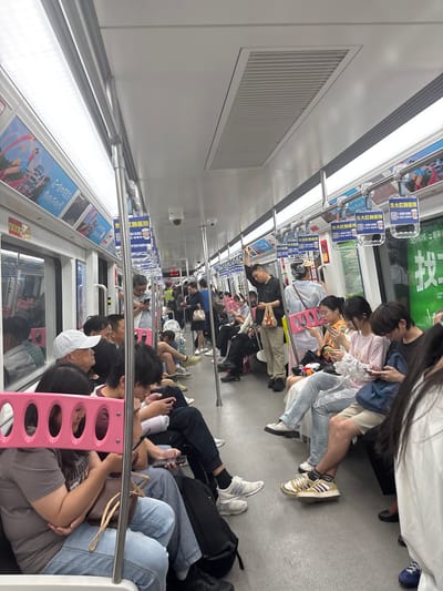 Here is a forensic description of the image:

**Scene:** The image captures a crowded subway car in what appears to be 龙溪街道, China.

**Environment:** The subway car features white interior panels with air vents along the ceiling. Overhead, there are advertisements, including signs for a medical institution.

**People:** The car is filled with passengers, many of whom are seated and occupy most of the visible space. Several people are holding and looking at their phones.

**Objects:** Key objects include cell phones, handbags, advertisements and subway handrails.