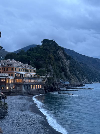 This image captures a coastal scene in Camogli, Italy, observed during dusk or dawn under an overcast, grey sky. On the left, a large, multi-story building with a light yellow facade and a tiled roof is prominently situated above a pebbled beach. The lower two levels of this building feature numerous illuminated arched windows and glass-enclosed sections, suggesting commercial or public spaces. A grey pebbled beach occupies the immediate foreground, with gentle white-crested waves breaking upon it from the deep blue-grey sea. To the right, steep, heavily vegetated cliffs rise sharply from the water, extending into the background where a prominent, tree-covered peak is visible, partially obscured by the low cloud cover. Smaller, multi-tiered structures are integrated into the cliff face further along the coastline, with some artificial structures or natural rock formations extending into the water. No individuals or specific human activities are visible.