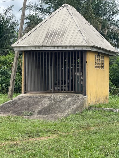 A small, yellow, concrete building with a pyramidal, metal roof sits in a grassy area, likely in a rural or suburban location. The building has a gated entrance made of metal bars. A concrete walkway leads up to the building. The building features a square latticework vent near the top. Trees and other vegetation surround the building. The location is Abraka, Nigeria.