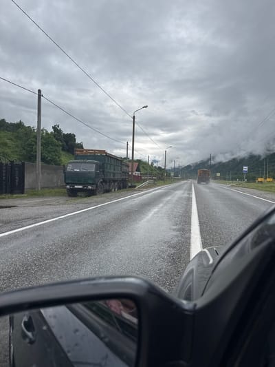 The image captures a rainy day on a road in Ново-Тебердинское сельское поселение, Russia. The scene is viewed from inside a vehicle, looking out through the windshield and side mirror. A wet asphalt road stretches ahead, marked by a white line. The sky is overcast with thick, gray clouds, suggesting a gloomy and possibly cool day. The road is lined with telephone poles and streetlights, with lush green trees and vegetation bordering the right side. In the distance, a truck with a yellow cargo bed is driving on the road. Closer to the foreground, a dark green truck with a large cargo container is parked on the side of the road, alongside a concrete barrier and a black metal fence. The side mirror of the vehicle shows a reflection of its interior, including what appears to be a person. A blue road sign indicating a bus stop is visible on the left side of the road. The license plate on the parked truck appears to be Russian. The overall impression is of a somber, wet day on a rural road.