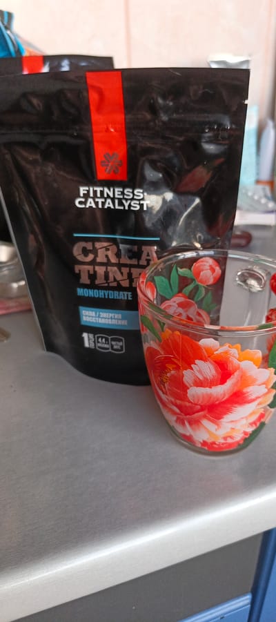This media file shows a close-up shot of a black pouch of "FITNESS CATALYST CREATINE MONOHYDRATE" next to a clear glass mug adorned with a floral pattern of red and pink peonies. The pouch has red and black branding with the brand name "FITNESS CATALYST" in white letters. Below the brand name, "CREATINE MONOHYDRATE" is prominently displayed in large, textured letters. Further down, text in Russian indicates "СИЛА / ЭНЕРГИЯ / ВОССТАНОВЛЕНИЕ" (Strength / Energy / Recovery). Icons below this text show "1 ПОРЦИЯ" (1 Serving), "44." with a droplet icon (likely indicating water or liquid), and "чистый вес" (net weight).
The glass mug is partially filled and exhibits a vibrant, hand-painted design of large peony flowers with green leaves. The items are placed on a brushed metal surface, likely a countertop. The background is blurred, showing a hint of a kitchen or bathroom setting with a beige tiled wall and some indistinct objects. The lighting appears to be natural, suggesting daytime. The scene is static, with no people visible, and the focus is on the product and the mug. The overall impression is of a preparation or post-workout scene. Given the location context of Tikhvin, Russia, the text on the pouch is in Russian, reinforcing this.