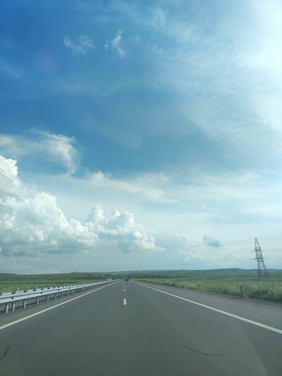 An asphalt road stretches into the distance under a vast blue sky filled with scattered white clouds. The road is marked with a white dashed line down the center and a solid white line along the right edge. To the left of the road, a silver guardrail runs parallel, separating it from a grassy verge. Beyond the verge, a flat, green landscape extends to a line of rolling hills in the background. A tall, lattice-work electricity pylon stands prominently on the right side of the road, silhouetted against the sky. Several other smaller poles and electrical wires are visible across the landscape. The scene appears to be a rural or semi-rural setting. No people are visible in the image.