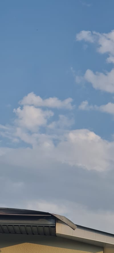 An upward-facing outdoor, daytime photograph shows a blue sky with scattered white clouds. In the upper third of the image, a small, dark object, possibly a bird or insect, is visible against the sky. The lower portion of the image features the roofline of a building with a dark metal roof and beige siding. The scene is a clear day with good visibility. The location is the city of Shaki, Azerbaijan.