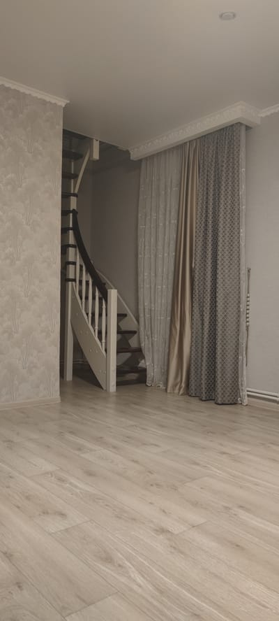 The image shows an interior scene with a spiral staircase on the left and curtains on the right. The floor is made of light-colored laminate flooring. The walls are wallpapered with a subtle pattern on the left and painted a neutral color on the right. The ceiling is white and has crown molding. The staircase is made of wood with white balusters and dark treads. The curtains are a mix of sheer and opaque fabric, with a gold-colored curtain in the center. There is a beaded decoration hanging on the right wall. The environment appears to be a residential interior, possibly a hallway or living area in a home in Икское Устье, Russia. No people or actions are depicted.