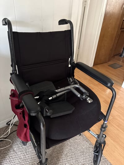 The image shows a black transport wheelchair with a maroon accessory bag attached to the back. The wheelchair is positioned on a carpeted floor next to a wooden floor. The background includes a wall with white horizontal panels and a section of wooden cabinetry. The wheelchair has black armrests, a black seat and back, and silver footplates. There is a label on the frame of the wheelchair that is partially visible. The scene appears to be indoors, in a home environment. There is no activity or event taking place, as the wheelchair is stationary. The lighting suggests it is daytime, and there are no indicators of weather or specific emotions. The location context is Bradenton, United States.